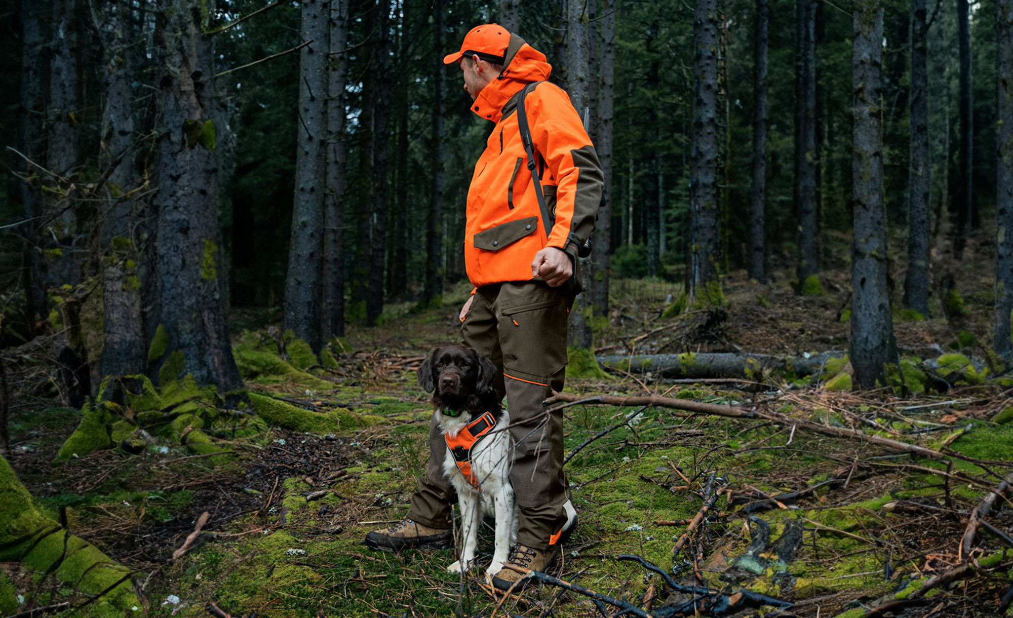 Ein Jger in orange-brauner Kleidung steht mit seinem Hund im moosigen Forst. Beide tragen auffllige Signalwesten fr bessere Sichtbarkeit.