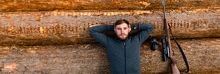 Ein Mann liegt entspannt mit verschrnkten Armen auf gestapelten Baumstmmen, neben ihm liegt ein Jagdgewehr mit Zielfernrohr.