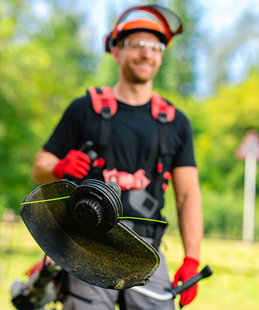 Mann mit Schutzhelm und Schutzbrille hlt einen Freischneider mit Fadenkopf in der Hand, im Hintergrund grne Natur.