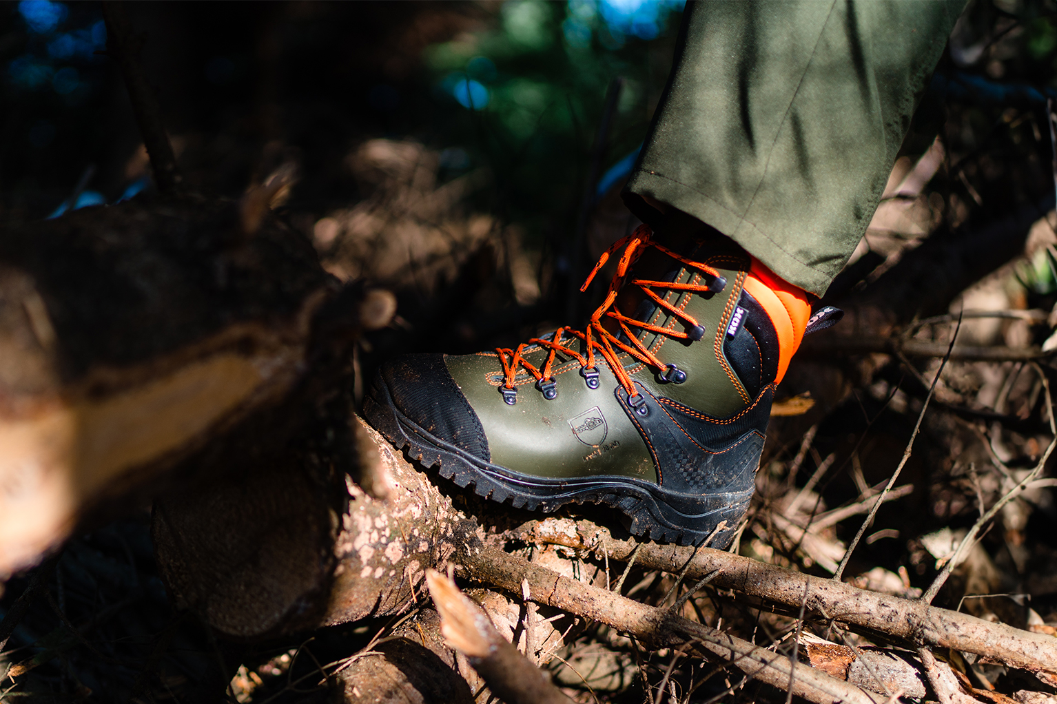 Ein robuster Forstschuh mit orangefarbenen Schn�rsenkeln steht auf �sten und Holz im Forst, die Profilsohle und der feste Schaft sind gut sichtbar.