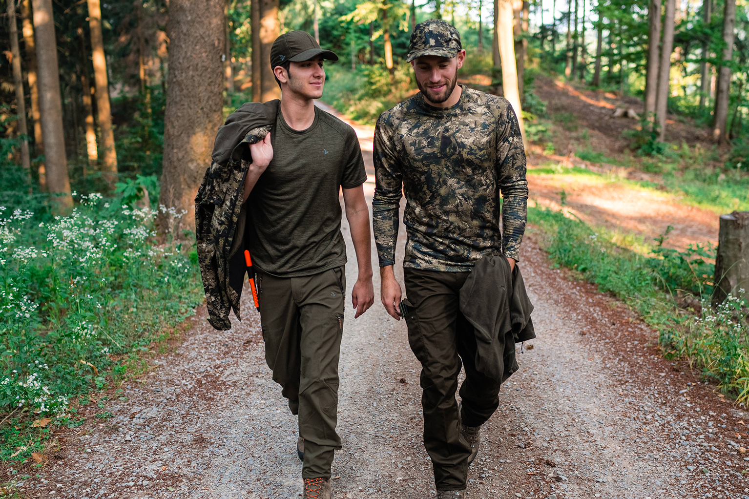 Zwei M�nner in funktionaler Outdoor- oder Jagdbekleidung gehen nebeneinander auf einem Waldweg; beide tragen Caps, Jacken in der Hand und unterhalten sich.
